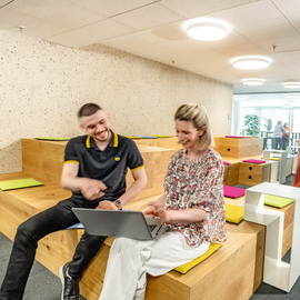 Zwei Mitarbeiter diskutieren zusammen am Laptop und sitzen im Headquarter auf einem Holzpalett