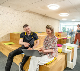 Zwei Mitarbeiter diskutieren zusammen am Laptop und sitzen im Headquarter auf einem Holzpalett
